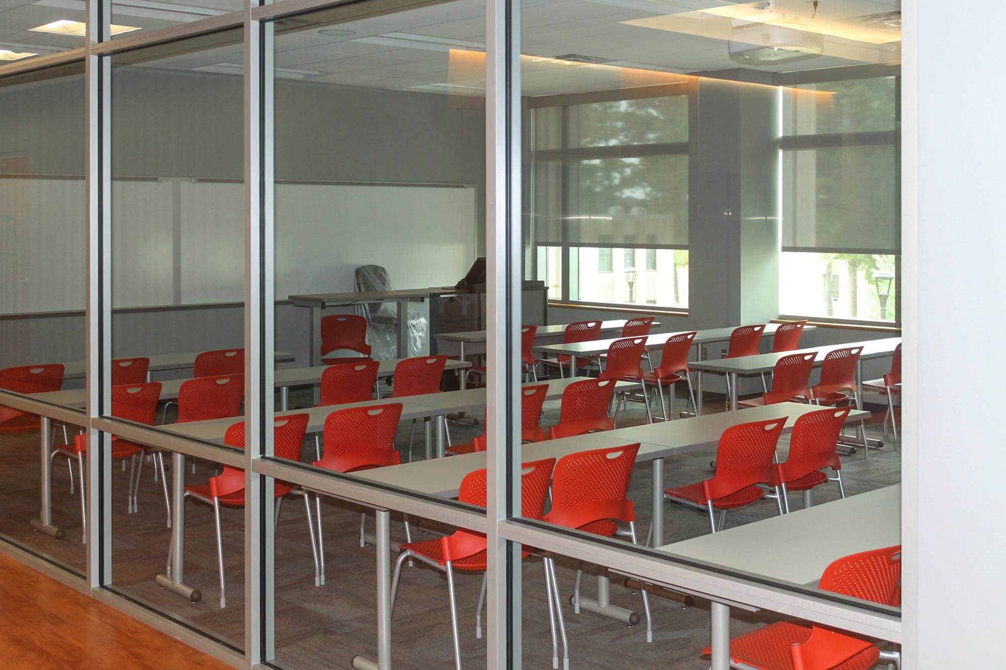 Classroom seen through glass partition furnished with long tables and orange chairs