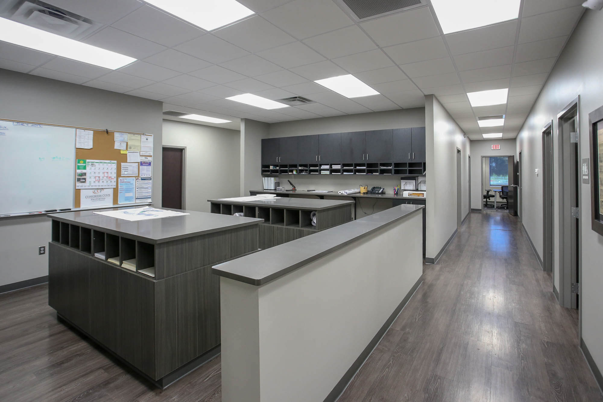 Interior shot of the Bailey Contractors, Inc. office workspace, featuring a central workstation with built-in storage cubbies, a bulletin board with notices, and a long hallway leading to private offices