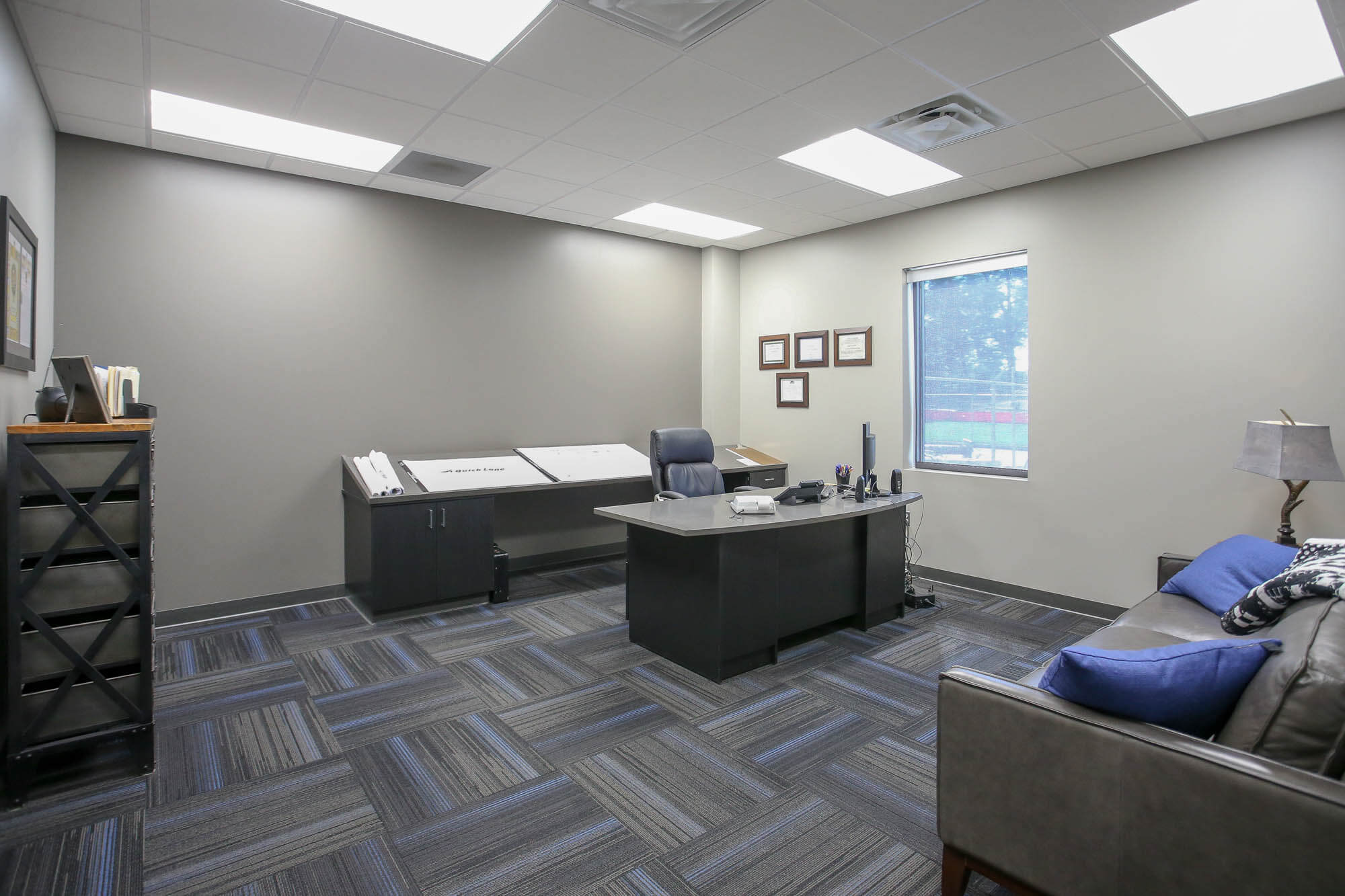 Interior shot of an executive office at Bailey Contractors, Inc., featuring a modern black desk with a leather office chair, a drafting table with architectural plans, and a seating area with a leather couch and blue accent pillows
