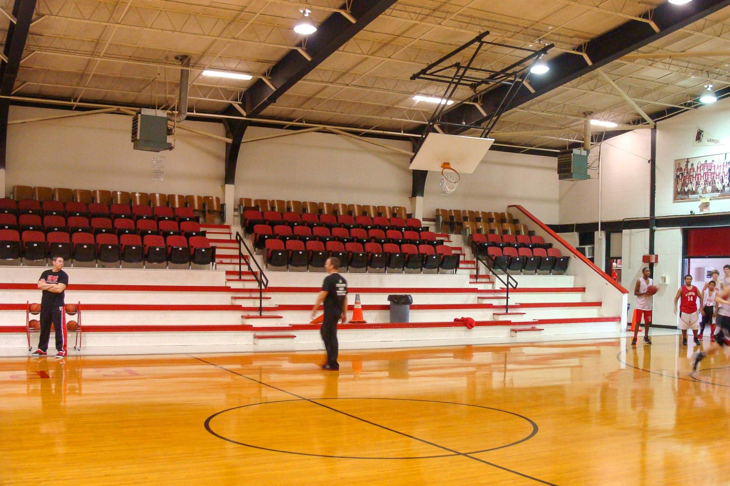 Gym interior with wood floor, red and black bleachers and students practicing basketball