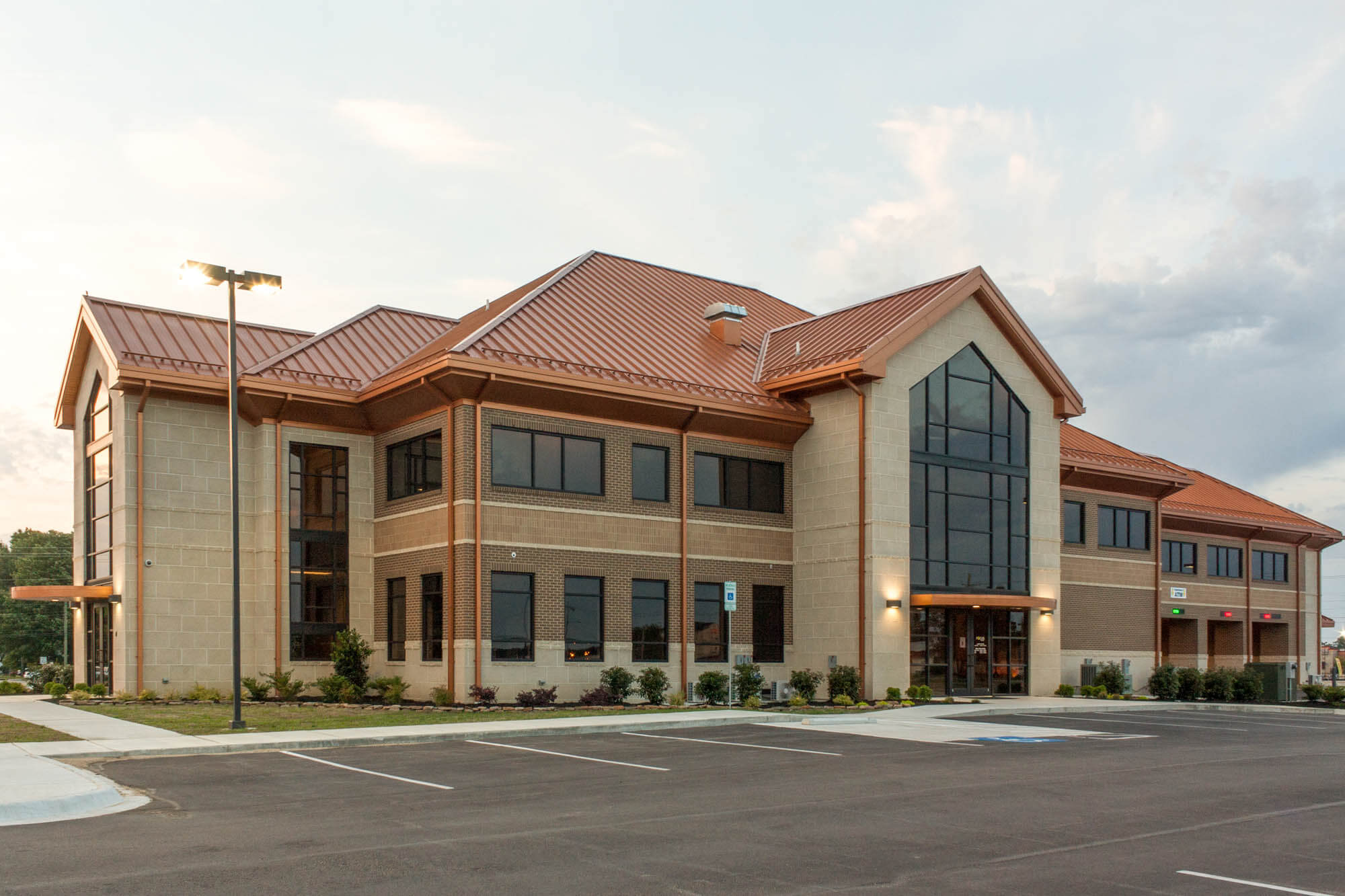 Angled exterior view of Focus Bank on Red Wolf Boulevard at dusk, showcasing a two-story brick and stone building with a copper-toned metal roof, tall central glass entryway, and an illuminated parking lot