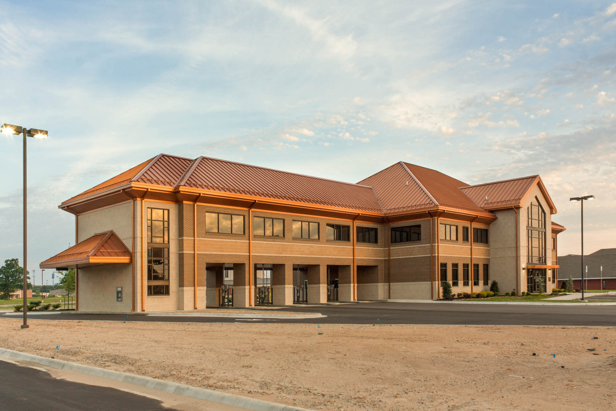 Angled exterior view of Focus Bank on Red Wolf Boulevard during golden hour, featuring a two-story brick and stone building with a copper-toned roof, drive-through lanes, and a large glass entryway