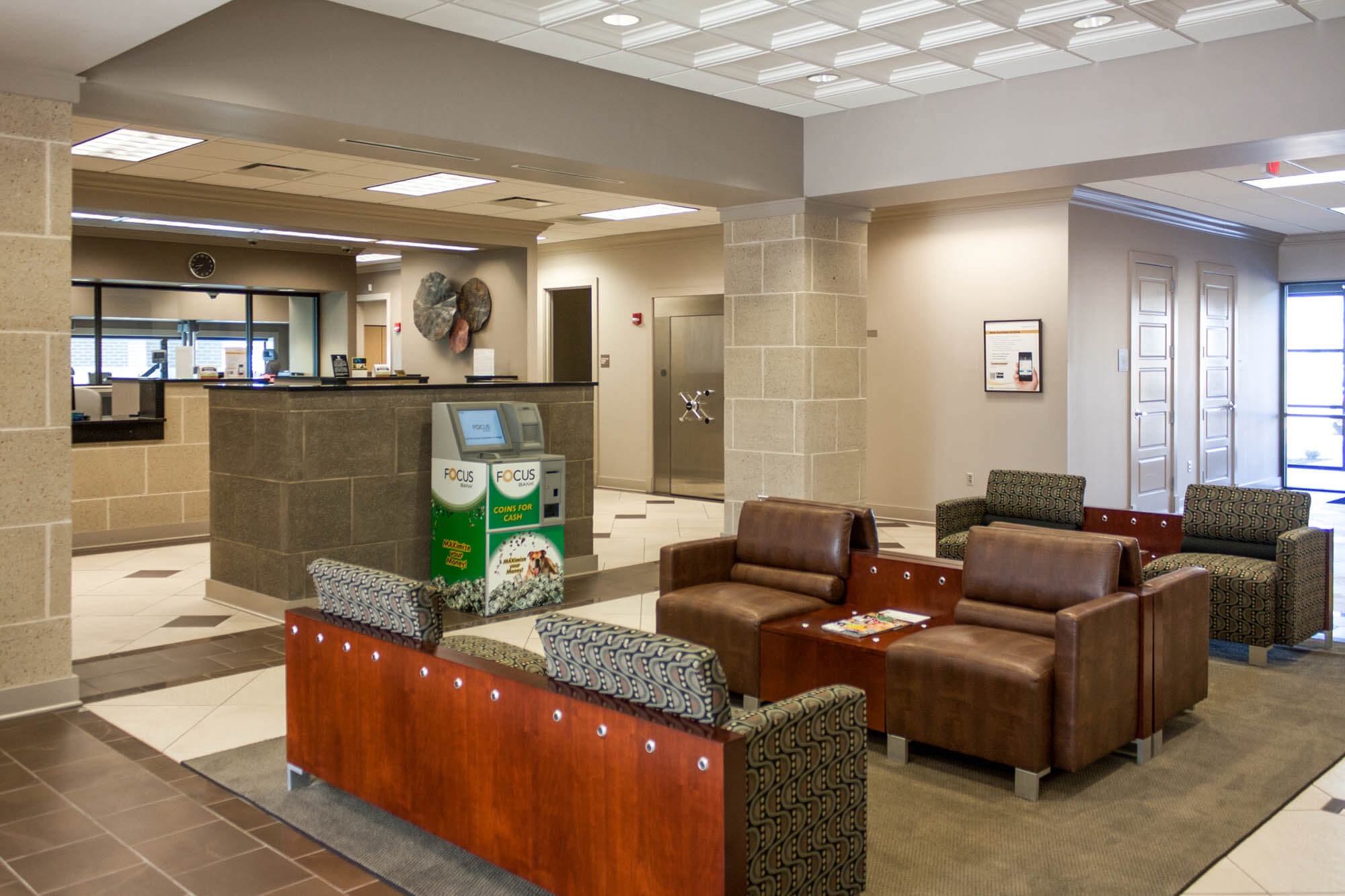 Interior view of the Focus Bank lobby on Red Wolf Boulevard, featuring a seating area with brown leather chairs and patterned armchairs, a coin machine kiosk, and the teller counter in the background with a vault door visible