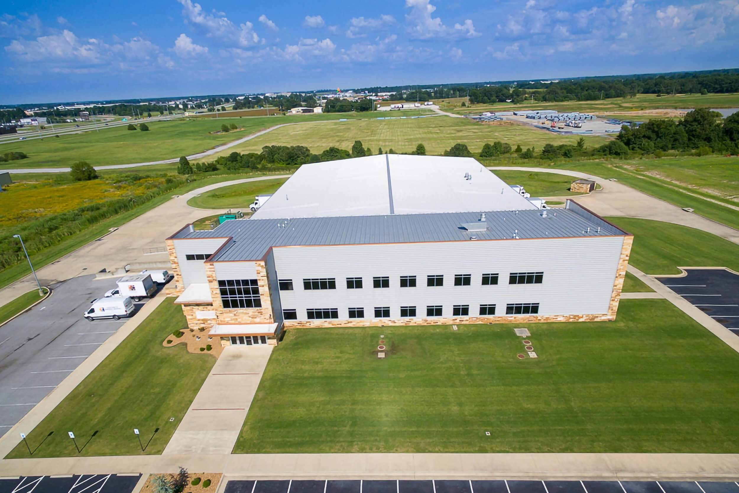 Aerial exterior drone shot taken from a high oblique angle, showcasing the front façade, and surrounding landscape, including parking areas and access roads