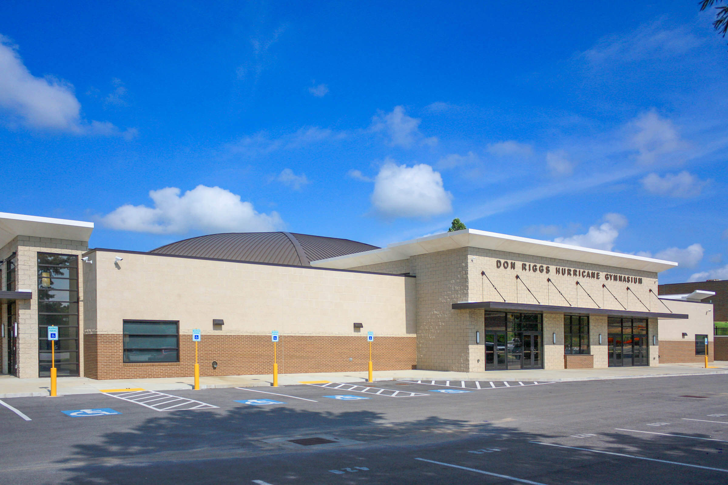 Exterior of the Don Riggs Hurricane Gymnasium showing cream masonry walls, curved roof, glass entry and adjacent parking spaces