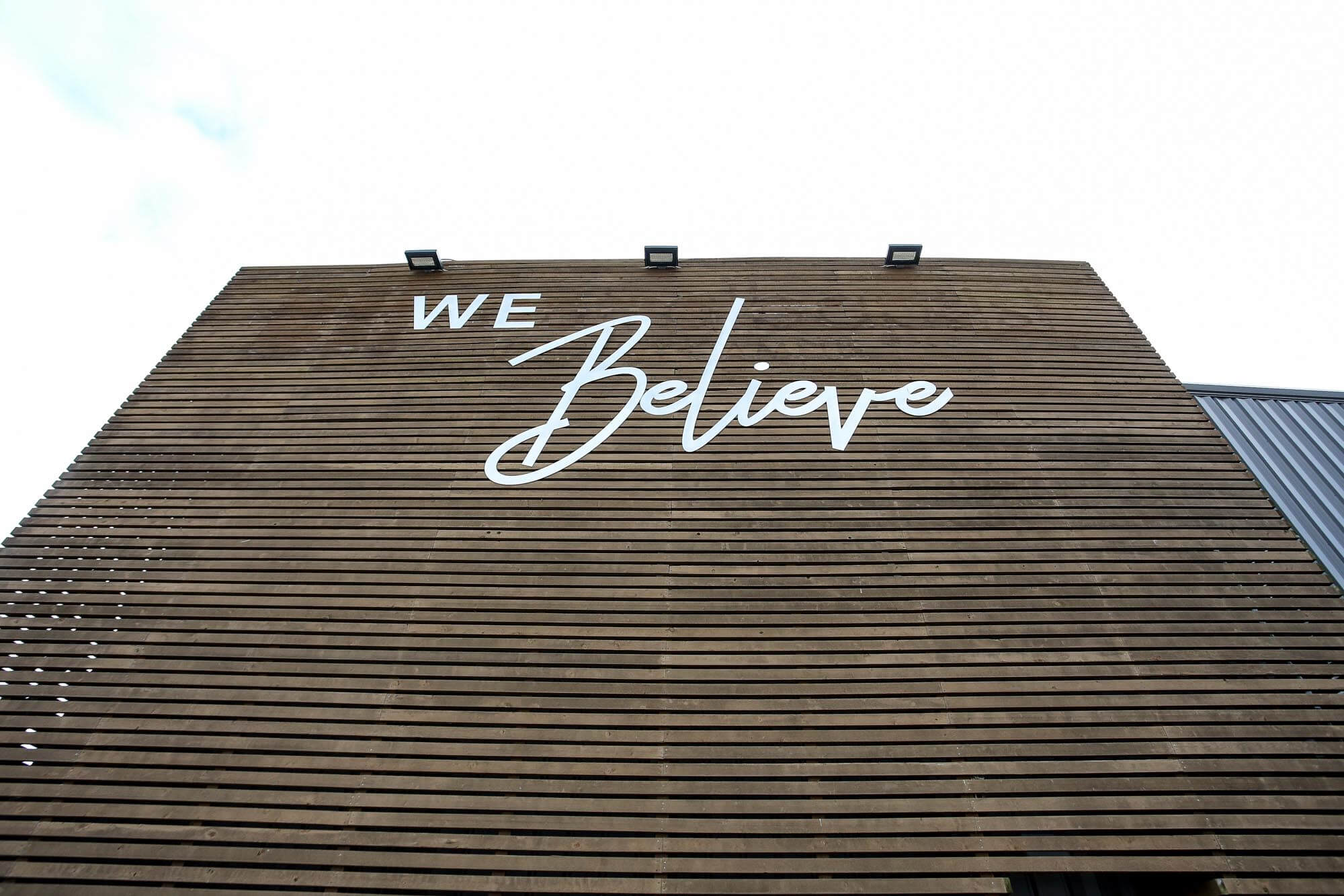 Close-up of Journey Baptist Church wood slat tower with large white 'We Believe' sign and mounted exterior lights