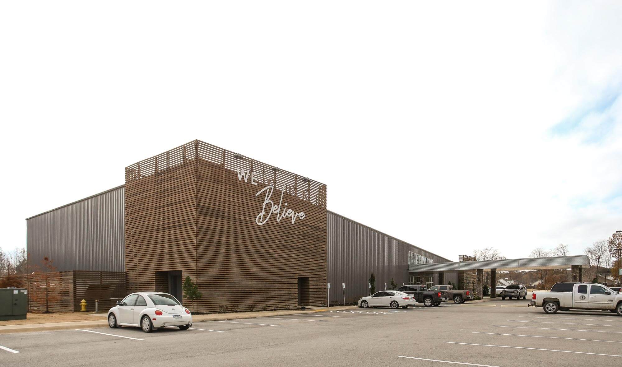 Exterior of Journey Baptist Church featuring modern design with wood slat tower, large 'We Believe' sign, metal siding, and covered entryway with cars parked in front