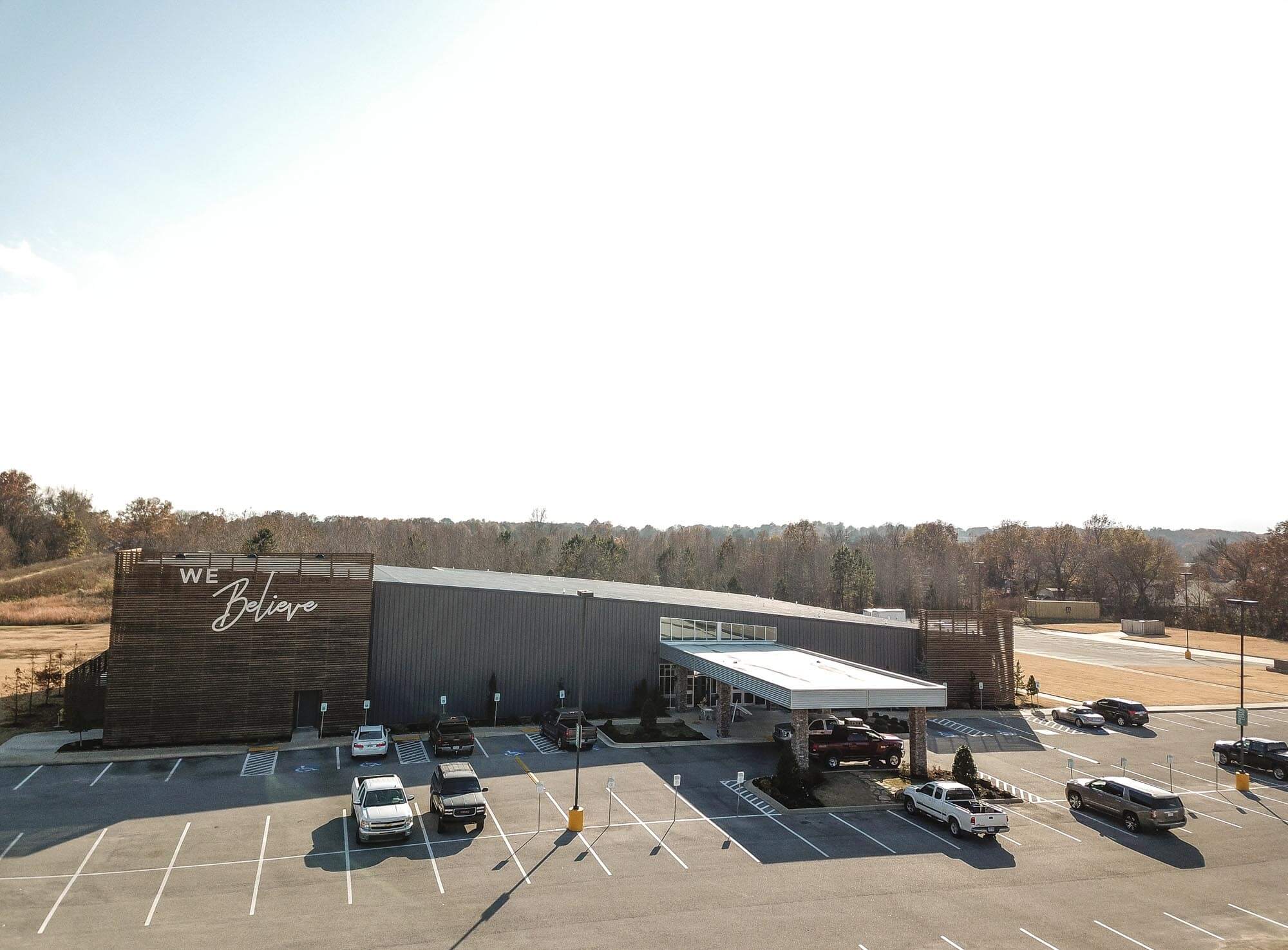 Aerial view of Journey Baptist Church with 'We Believe' sign on the wood slat tower, large gray exterior, parking lot, and tree line in the background