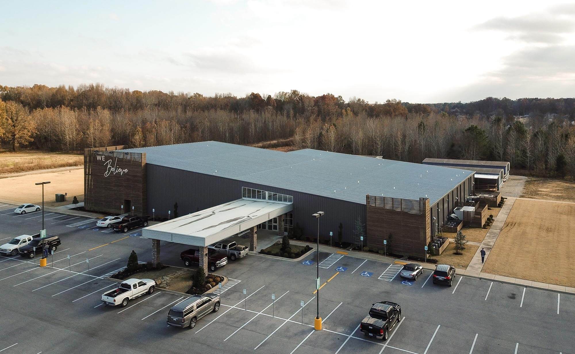 Wide aerial view of Journey Baptist Church building with gray roof, wood slat towers, front awning entrance, parking lot, and wooded backdrop