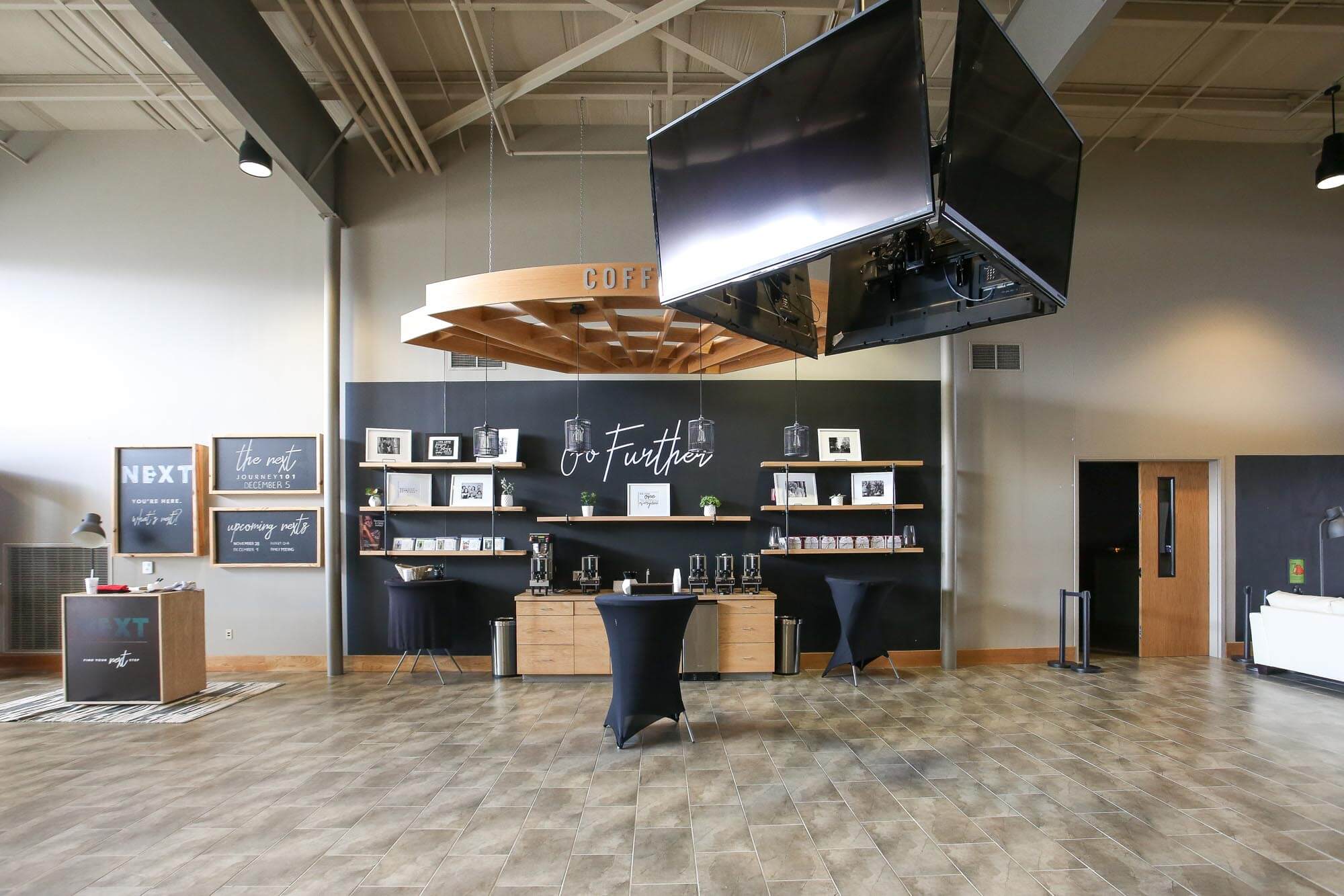 Journey Baptist Church interior coffee bar with hanging wood feature, black accent wall with Go Further sign, shelves, self-serve coffee station, and four TVs suspended from the ceiling in a square rig