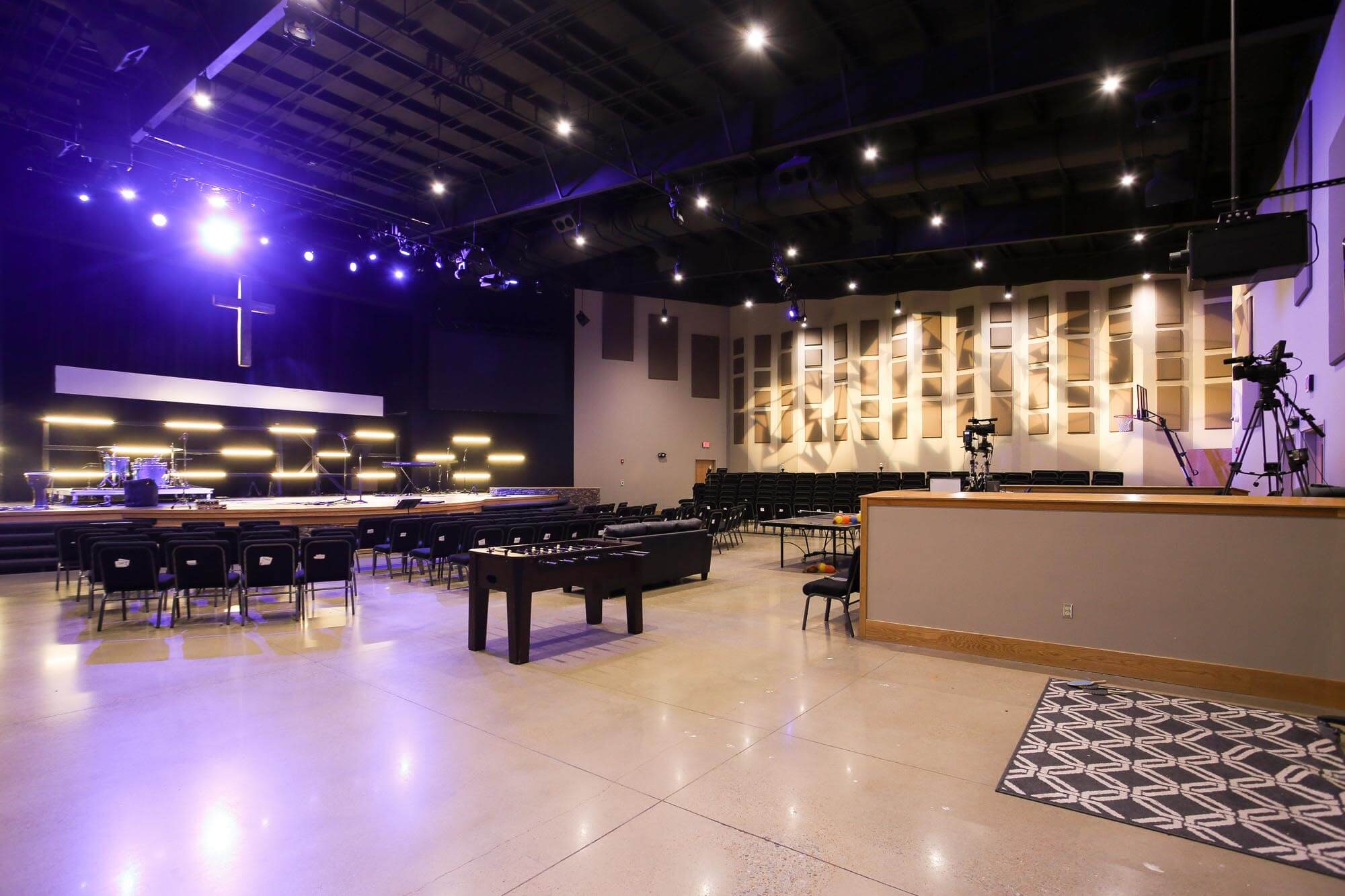Journey Baptist Church sanctuary with stage lighting, illuminated cross on back wall, drum set and instruments on stage, rows of chairs, sound cameras on tripods, acoustic panels on walls, and a foosball table in the seating area