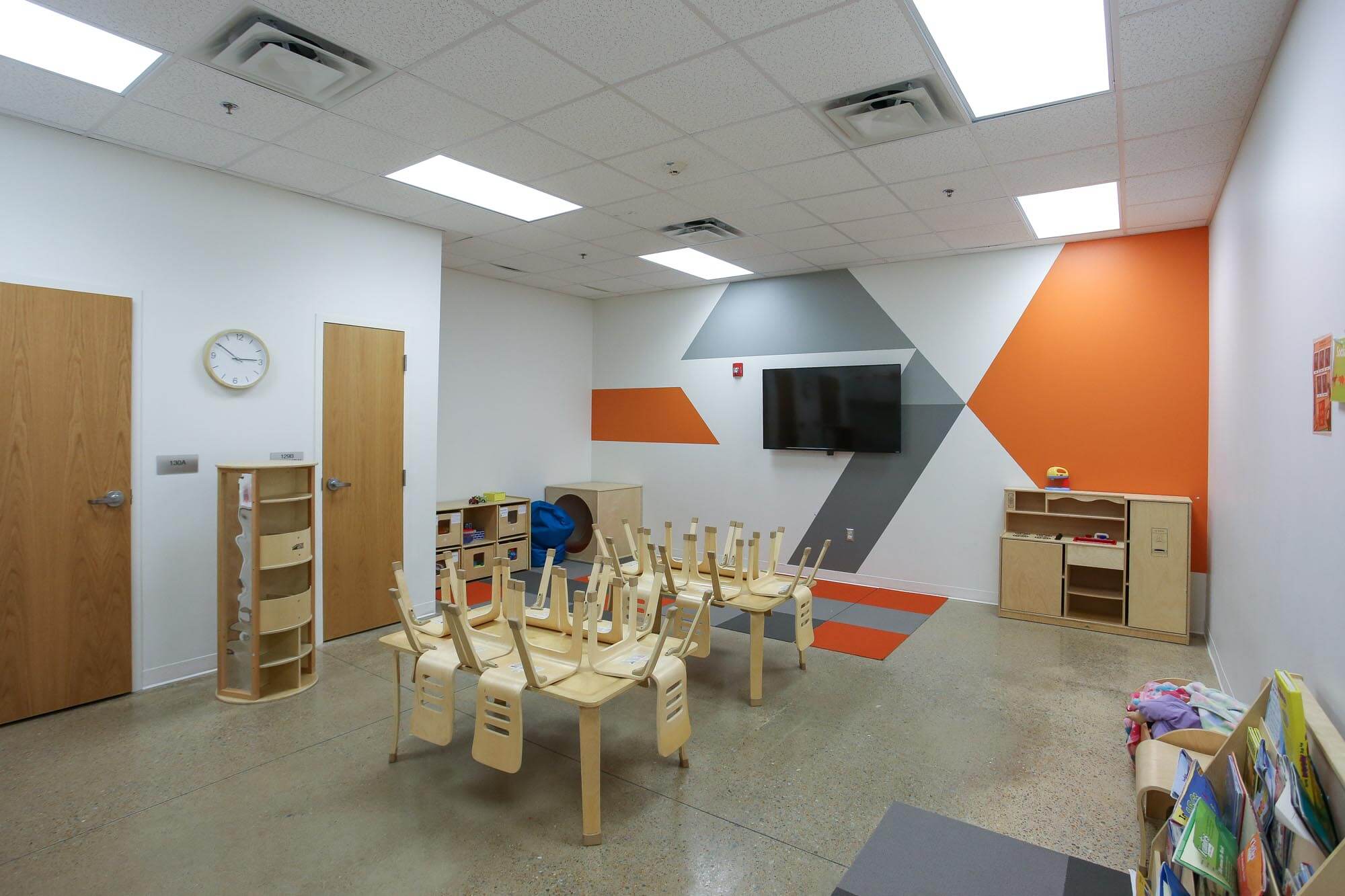 Journey Baptist Church kids classroom with colorful geometric wall design in orange and gray, mounted TV, small wooden tables and chairs stacked upside down, cubby shelves, and children’s books in a reading corner