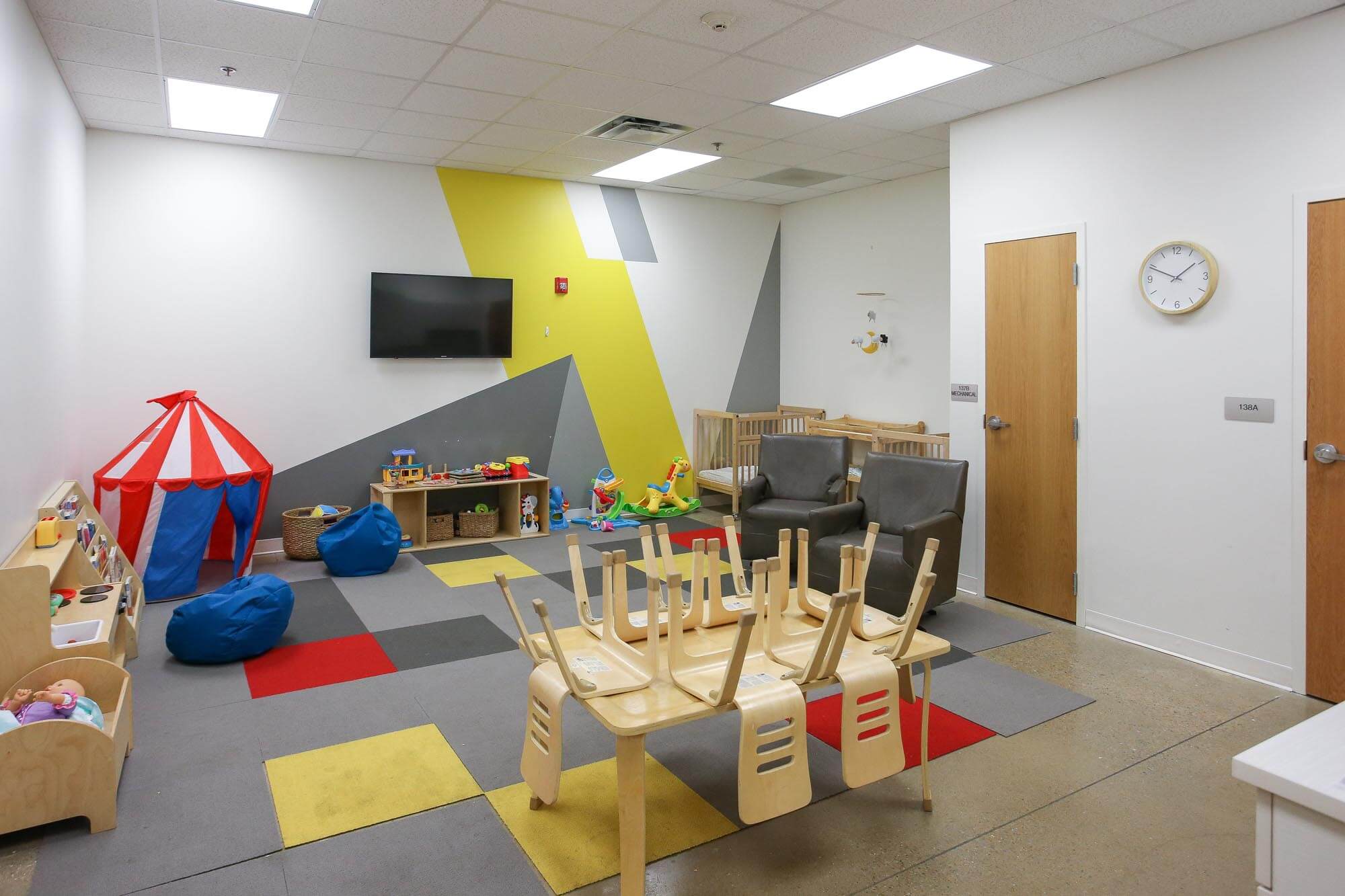 Journey Baptist Church nursery with colorful play mats, red and white play tent, toys, wooden cribs, beanbag chairs, and wall-mounted TV