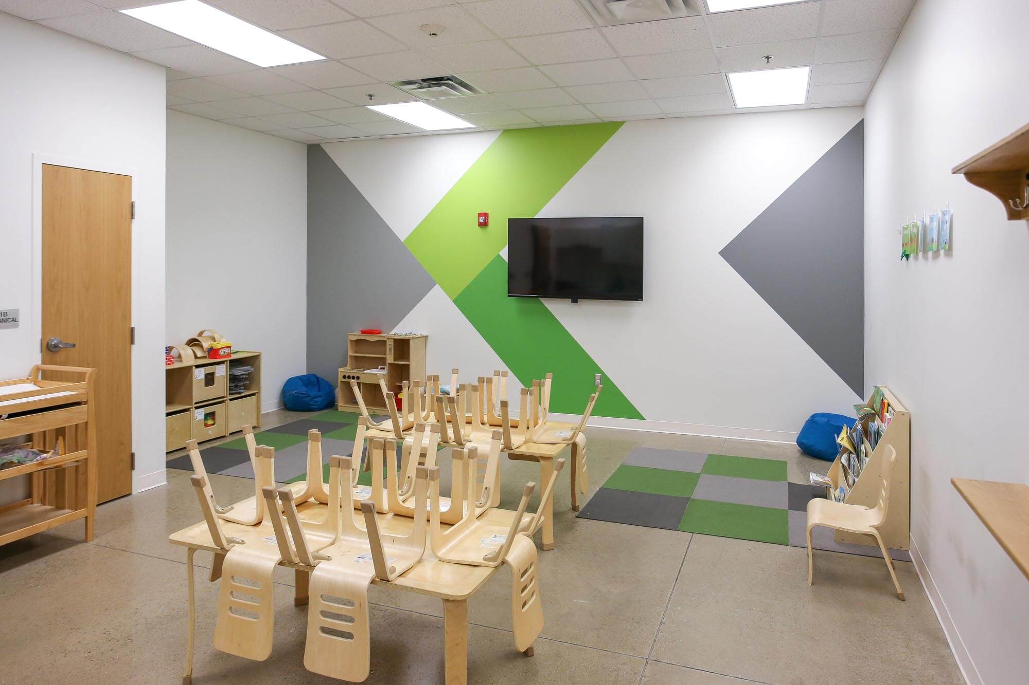 Journey Baptist Church kids classroom with colorful green and gray geometric wall design, small tables with chairs stacked upside down, wall-mounted TV, bookshelves, and play area