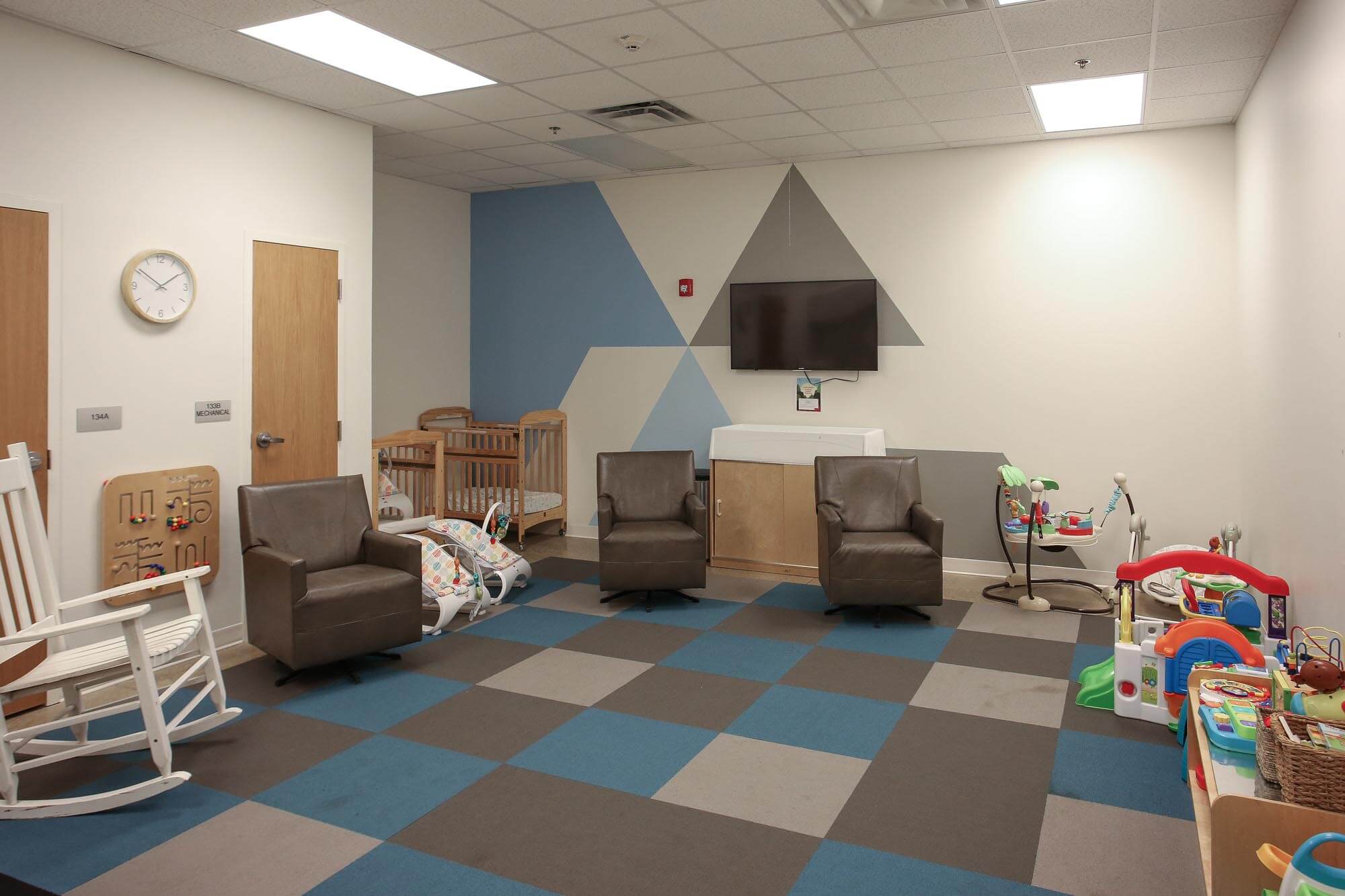 Journey Baptist Church nursery with blue and gray patterned floor, geometric blue and gray wall design, cribs, rocking chair, leather chairs, play toys, and wall-mounted TV