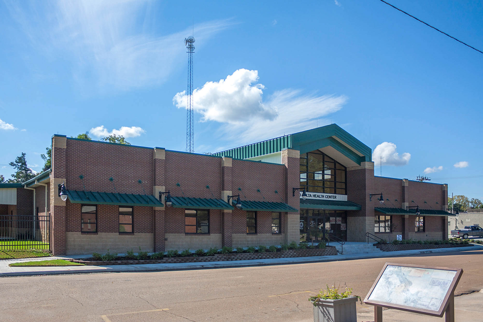 Front exterior of the brick health center with green roof and gabled glass entrance, plus walkway, signage and signboard