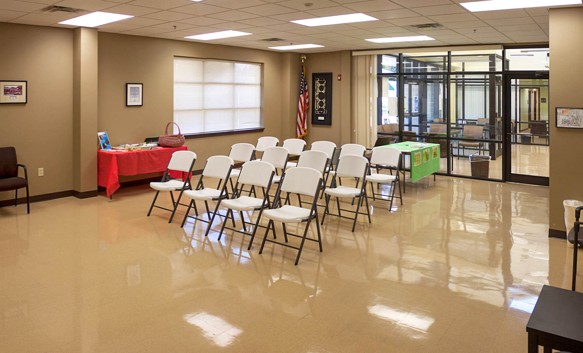Multi‑purpose meeting room featuring rows of folding chairs, an American flag and glass partition to the waiting area