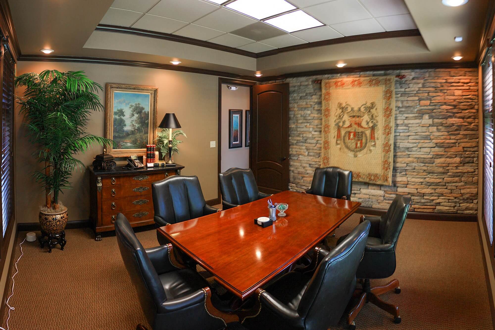 Conference room with polished wooden table, black leather chairs, stone accent wall, decorative crest tapestry and potted plant