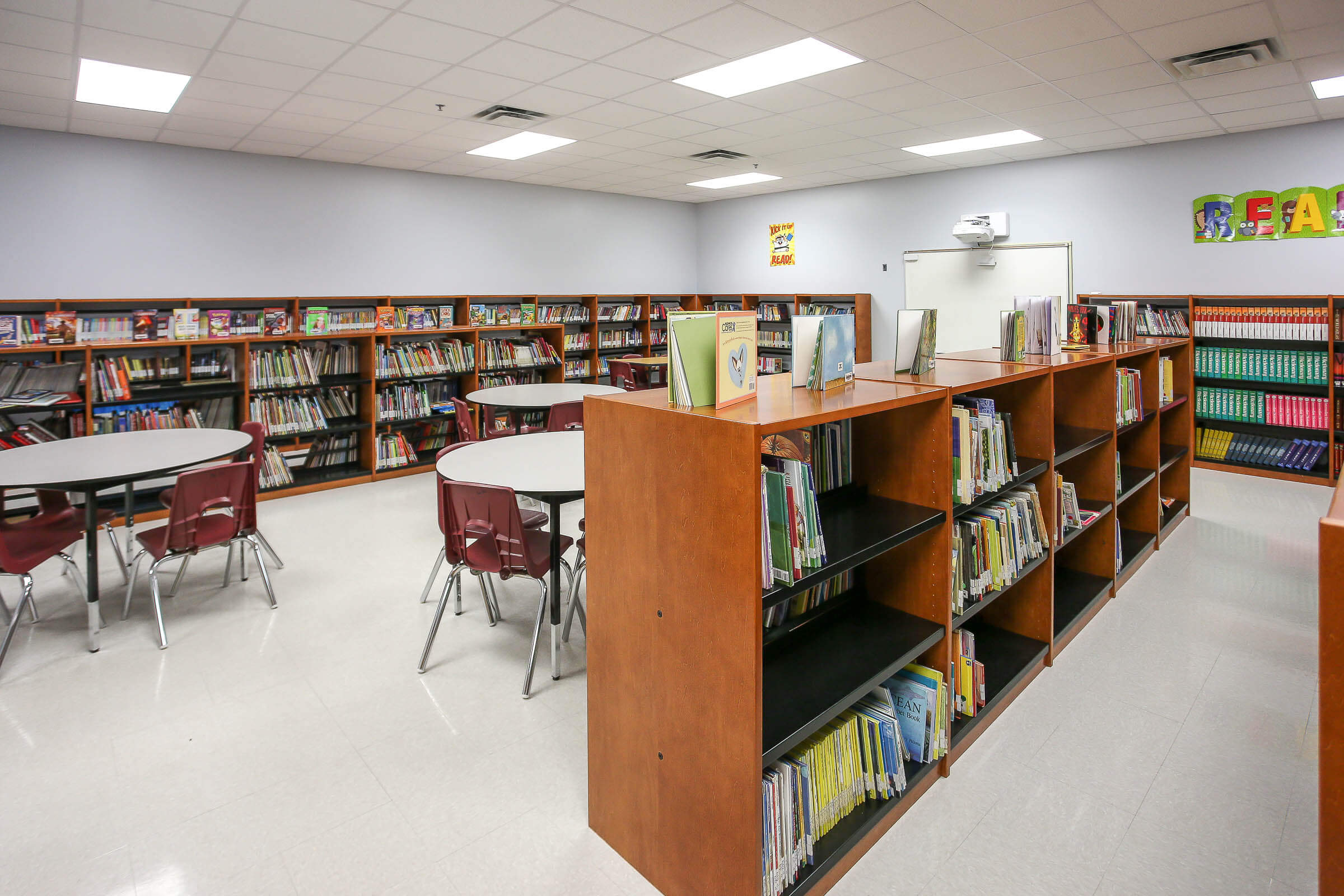 Library interior featuring bookshelves, round tables, projector, grey tile floor and walls decorated with posters