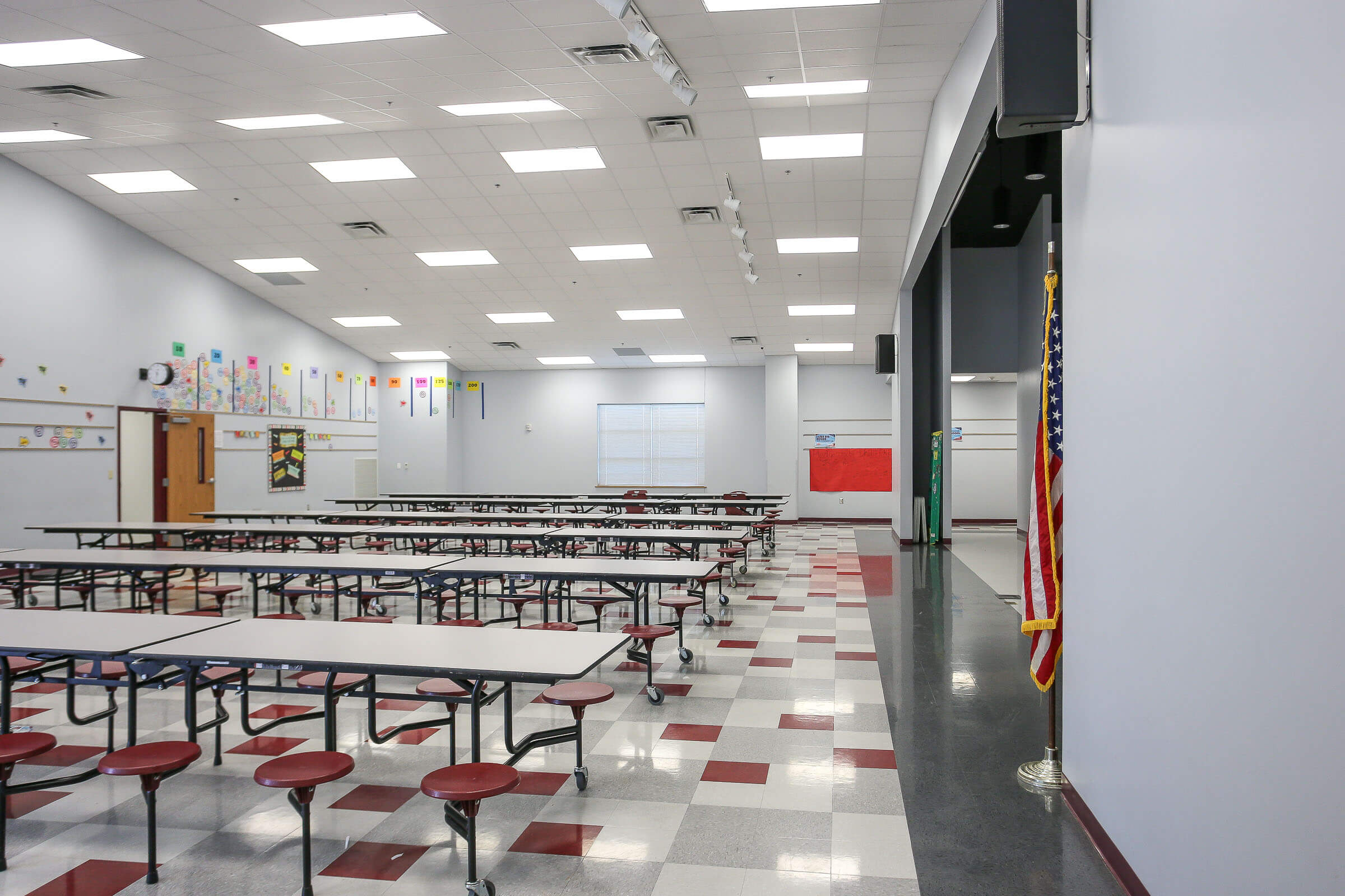 Empty cafeteria with multiple tables and red stools, grey walls, stage area and American flag
