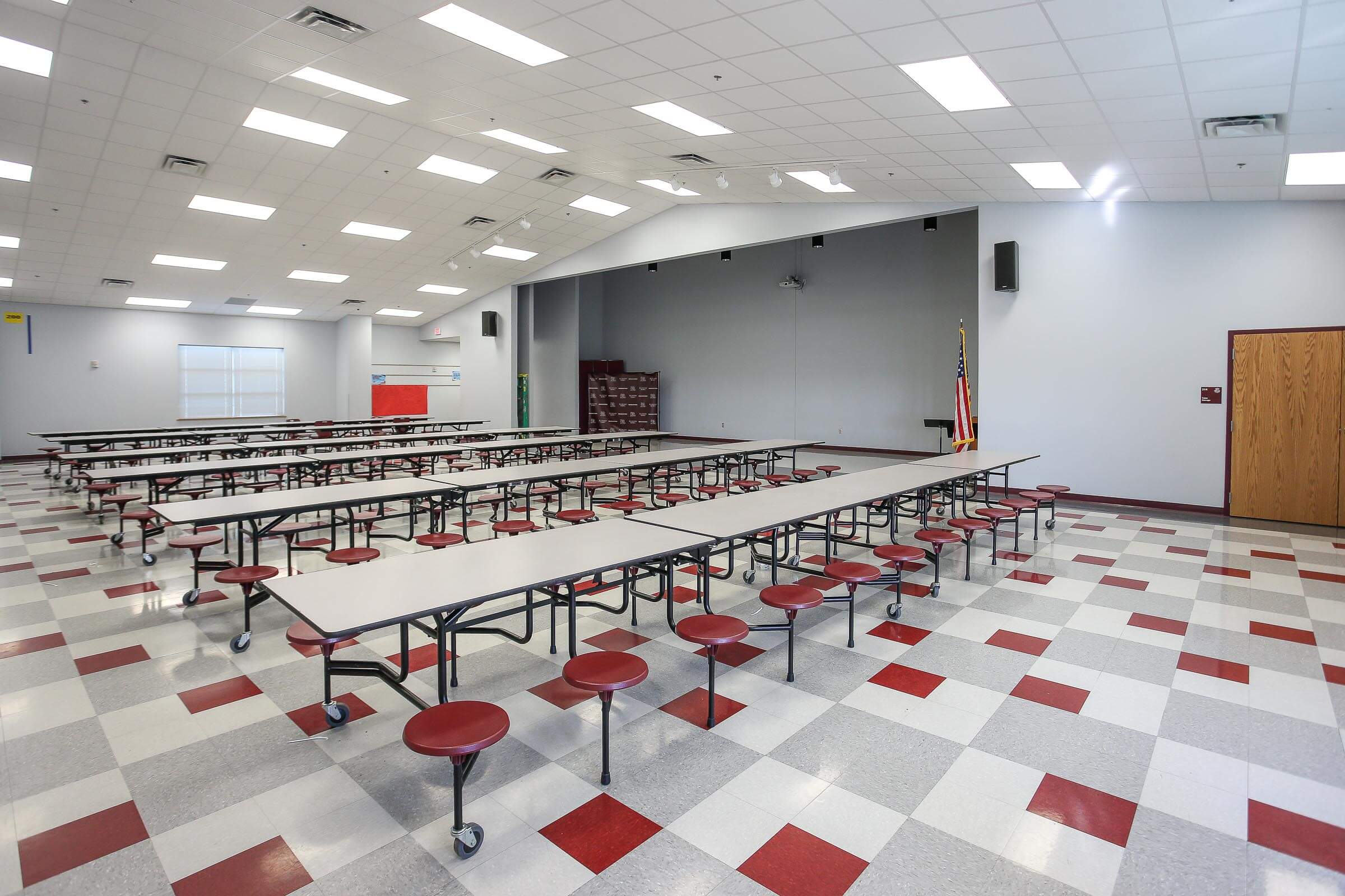Empty cafeteria with multiple tables and red stools, grey walls, stage area and American flag, view from stage area