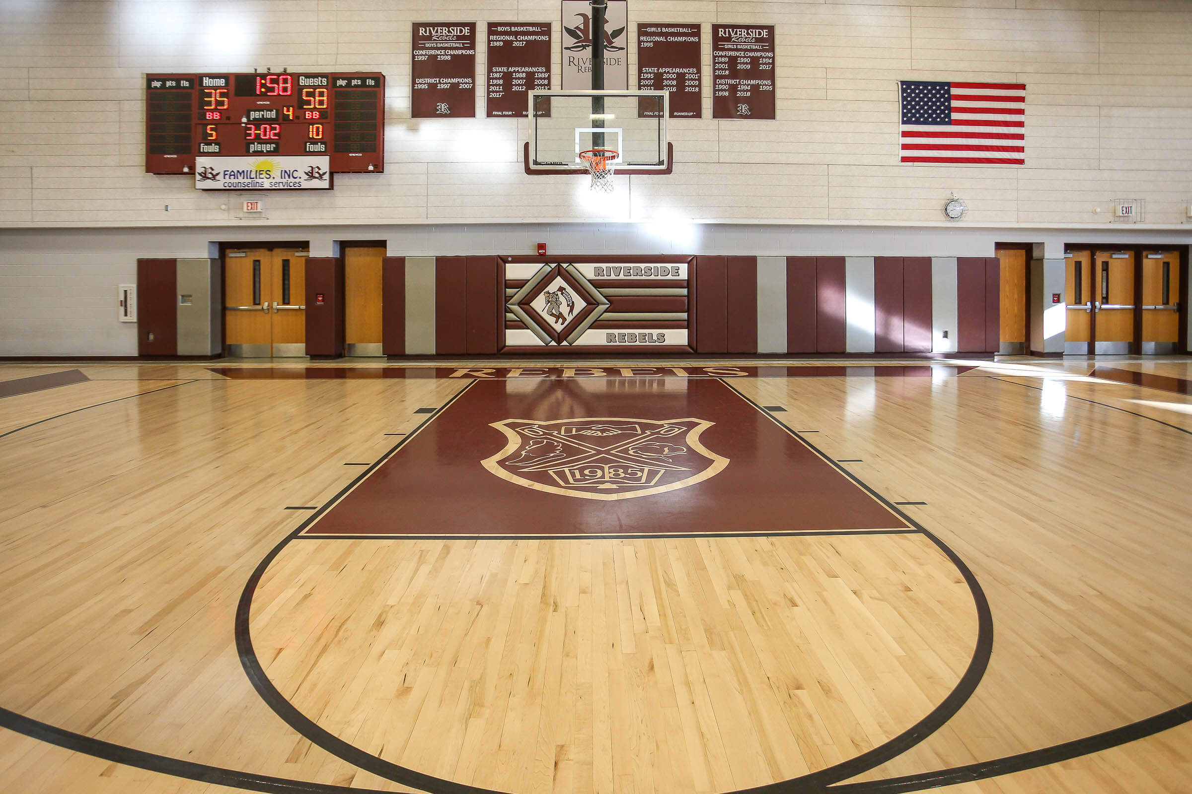 Gymnasium view focusing on the scoreboard, basketball goal and center crest on the floor