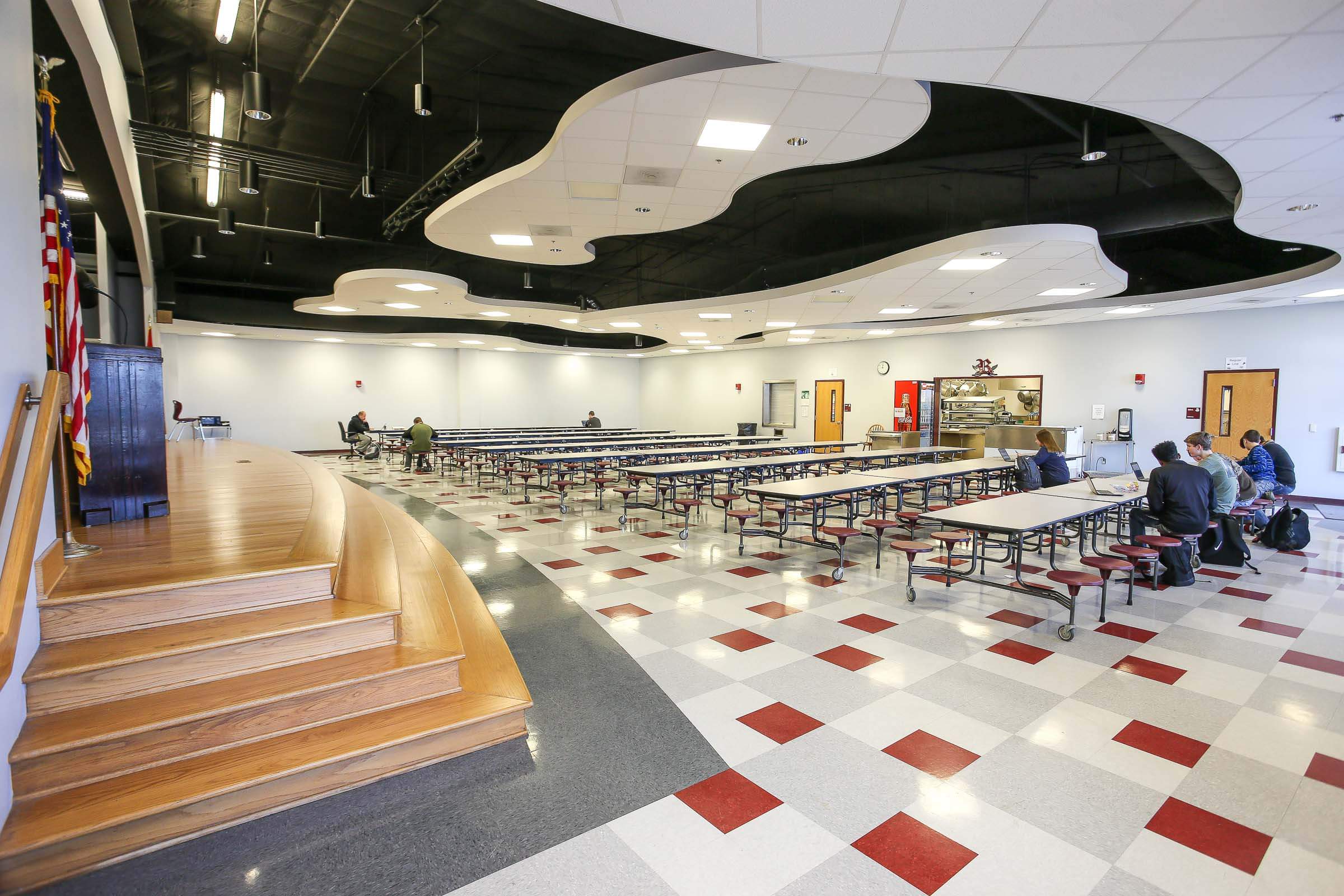 Cafeteria with built-in stage, wavy black-and-white ceiling panels and tables with attached stools