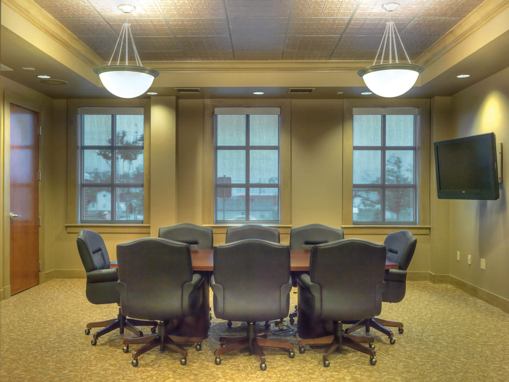 Conference room featuring a long table, black rolling chairs, windows with shades, pendant lights and wall-mounted television