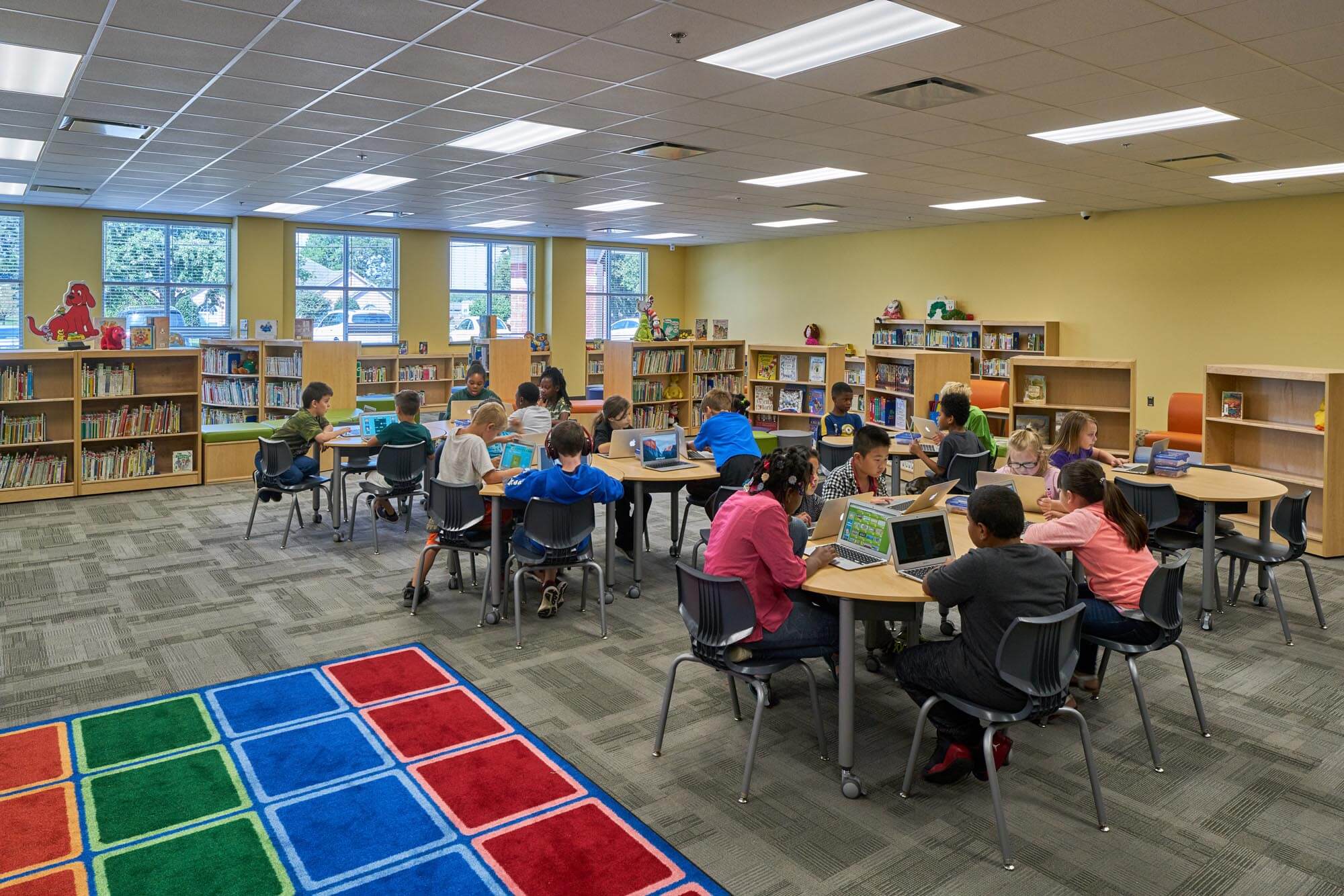 Elementary school library with students at round tables using laptops, surrounded by bookshelves