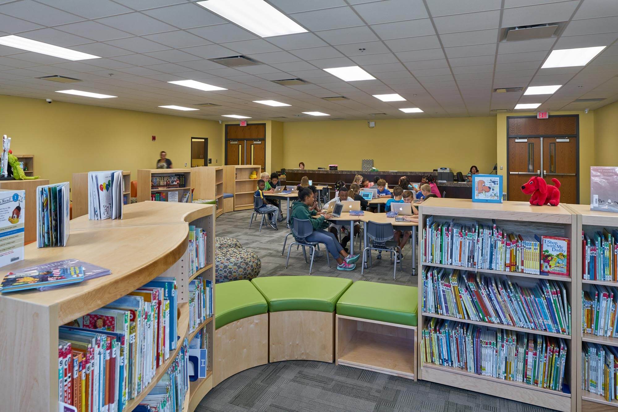 Students working on laptops at tables in a school library with bookshelves and seating in foreground