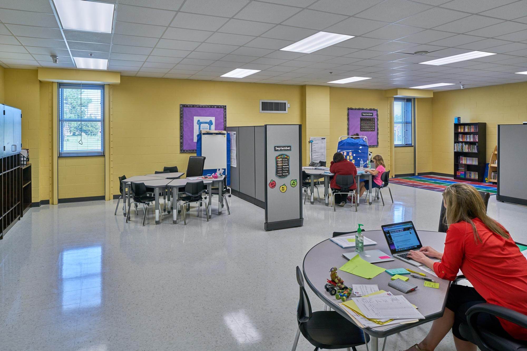 Teacher working on laptop at desk with small student groups in a classroom with partitions