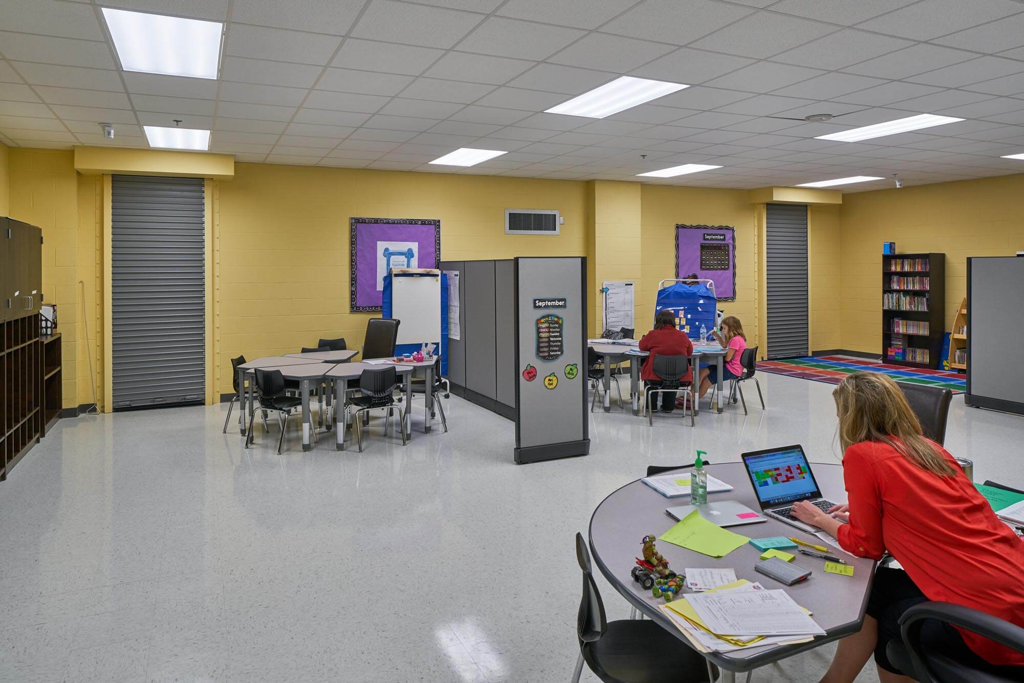 Teacher working at desk with laptop while small student groups study in classroom with partitions with blinds pulled down