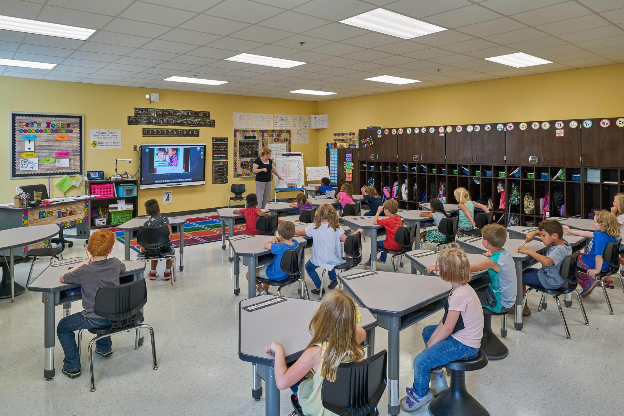 Elementary teacher leading lesson while students sit at desks in classroom with cubbies