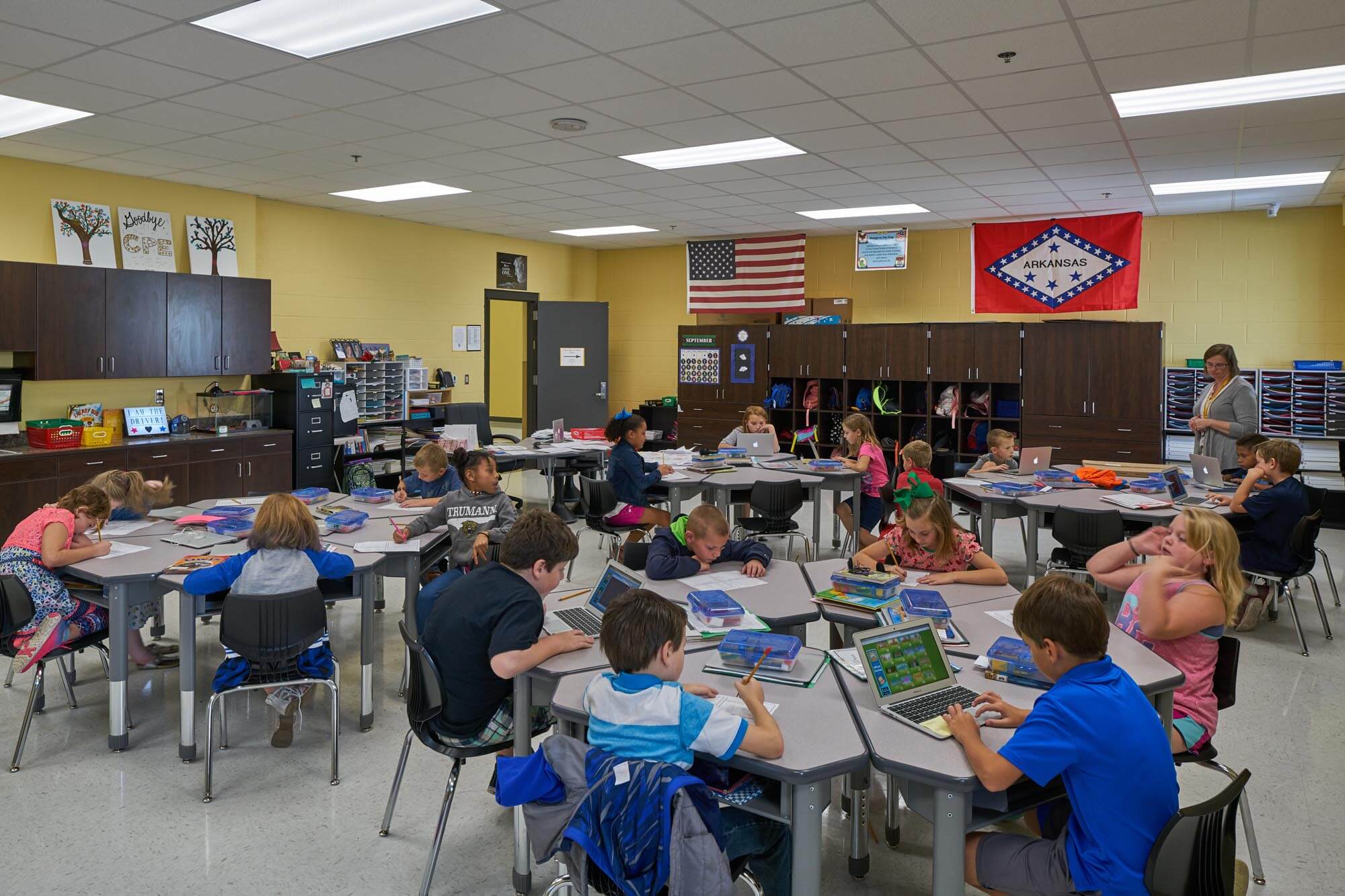 Elementary students working at tables with laptops and notebooks while teacher observes in classroom with Arkansas flag