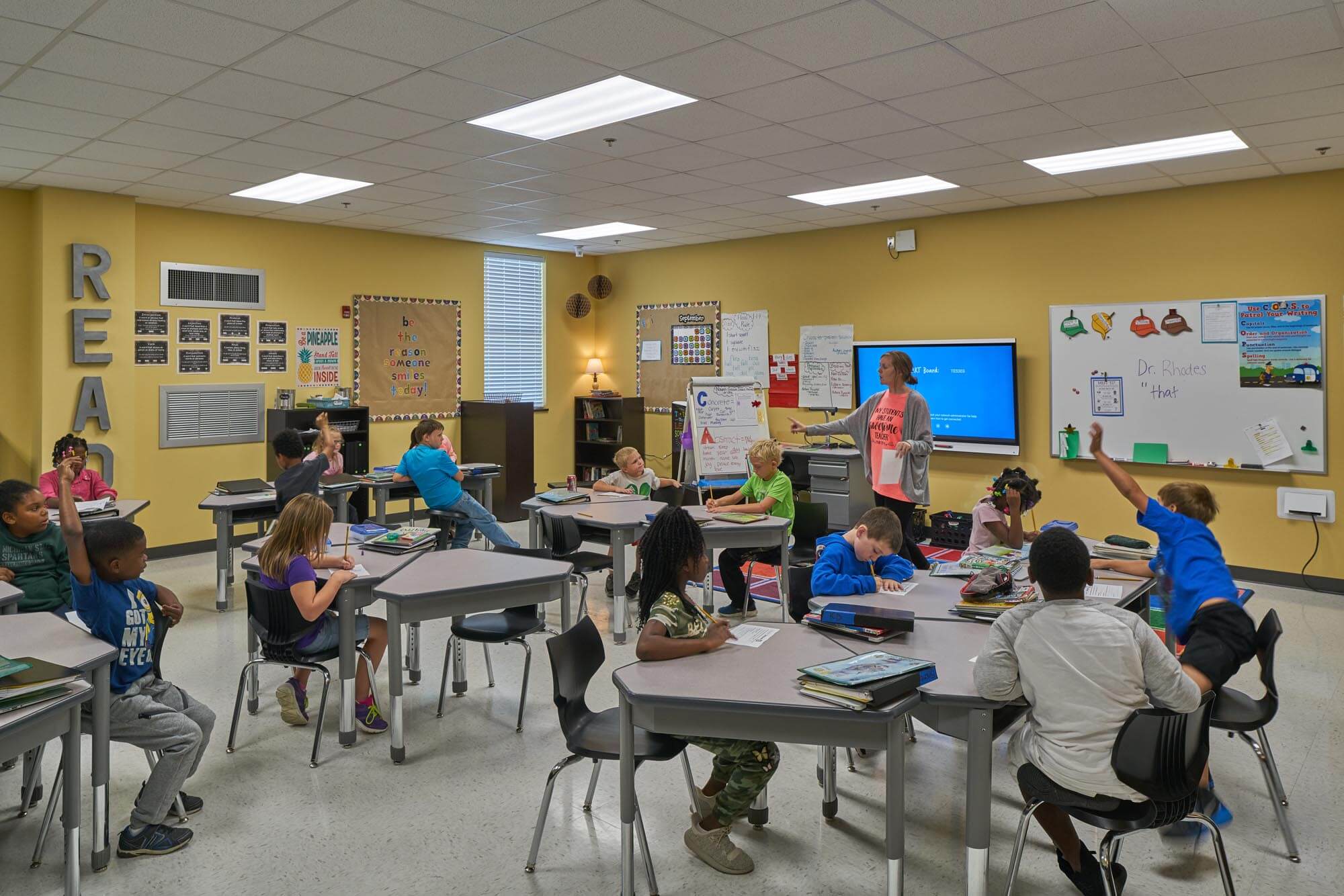 Elementary classroom with teacher leading lesson as students sit at tables, some raising hands