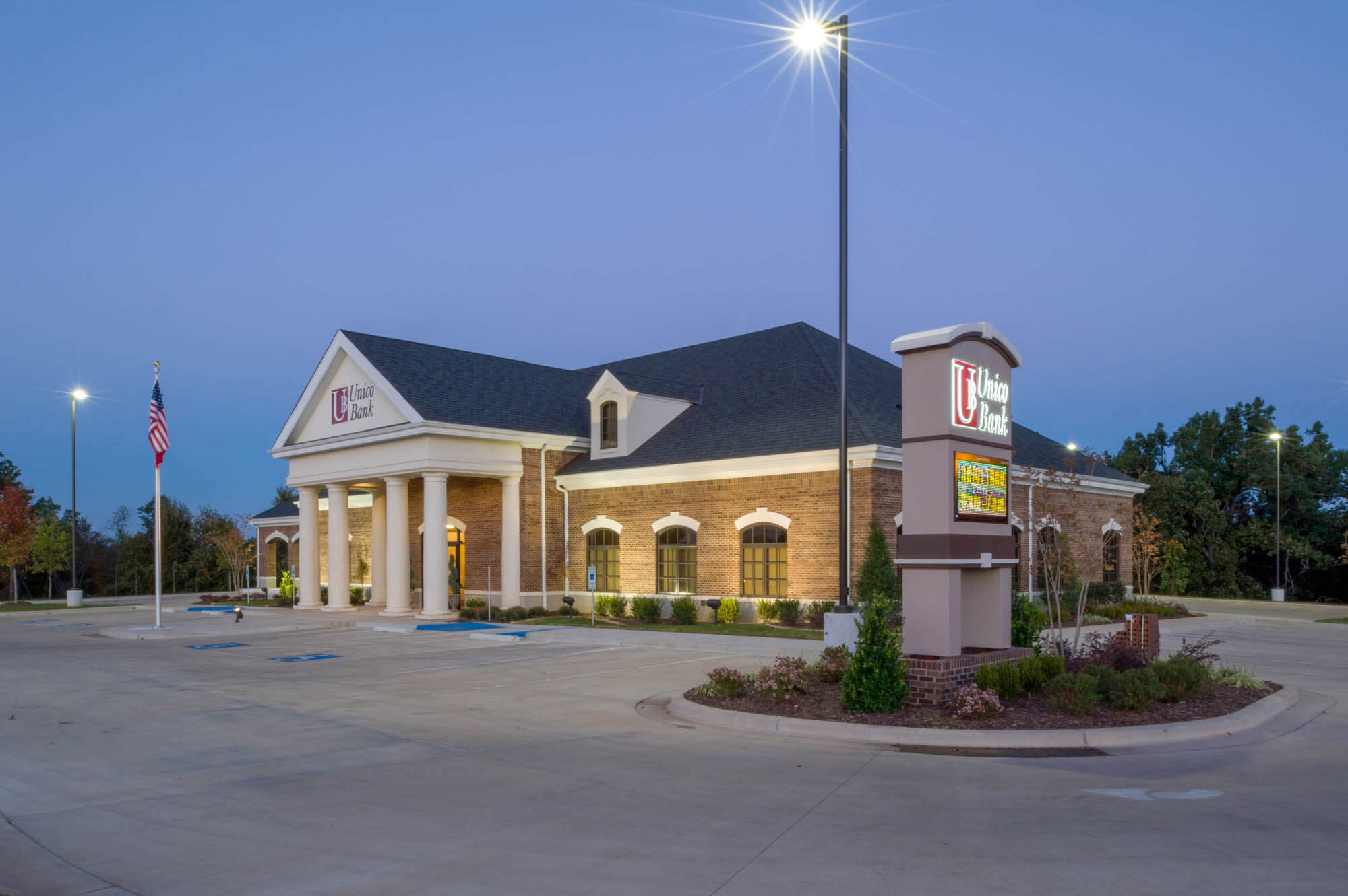 Twilight exterior of Unico Bank with brick facade, columned entrance, illuminated sign, lampposts and an American flag
