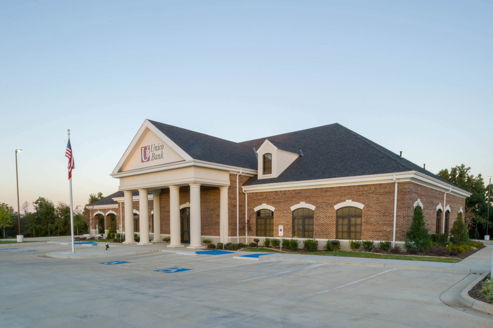 Angled exterior showing the bank’s dormer roof, columned porch and landscaped parking area with trees and shrubs