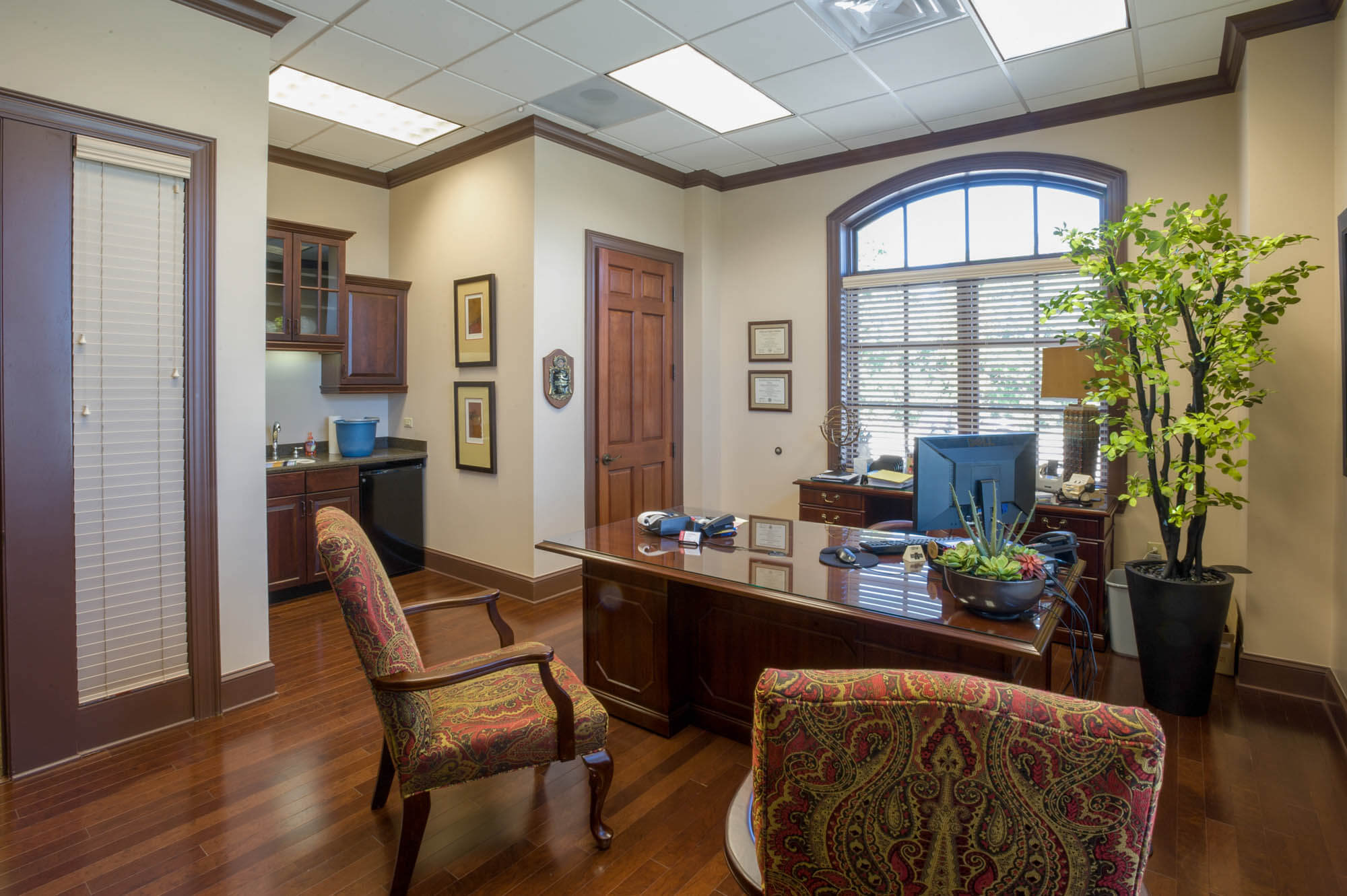 Private office furnished with wood desk, paisley upholstered chairs, arched window and kitchenette