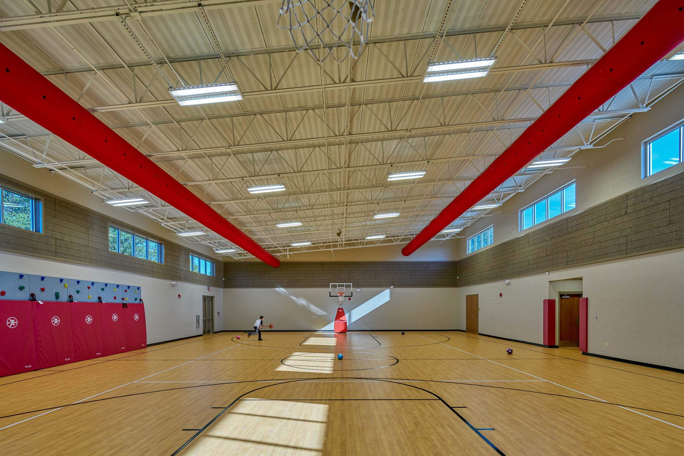 Gymnasium with wood floor, red duct along the ceiling, climbing wall and children playing with balls